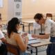 Mulheres em seção eleitoral com cabine de votação 'ELEITORAL'. Uma mesária auxilia eleitora com documentos