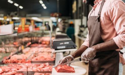 Açougueiro com avental e luvas corta carne vermelha em tábua, com balcão de açougue ao fundo