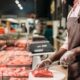 Açougueiro com avental e luvas corta carne vermelha em tábua, com balcão de açougue ao fundo