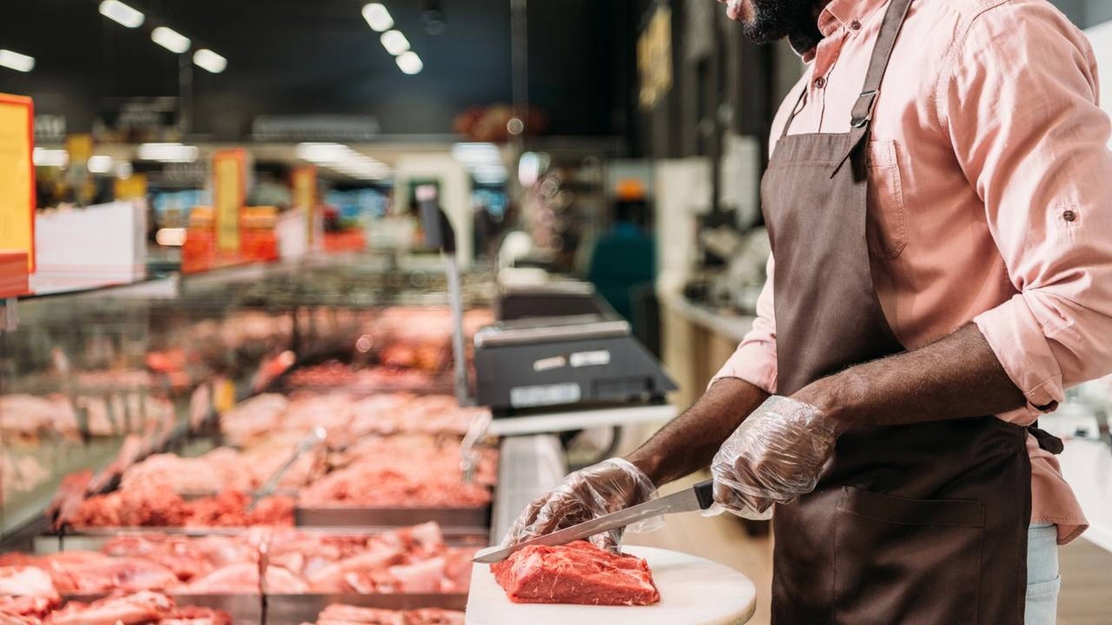 Açougueiro com avental e luvas corta carne vermelha em tábua, com balcão de açougue ao fundo