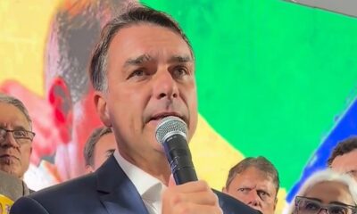 Homem de terno escuro com microfone, fundo com cores da bandeira do Brasil, outras pessoas ao redor