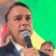 Homem de terno escuro com microfone, fundo com cores da bandeira do Brasil, outras pessoas ao redor