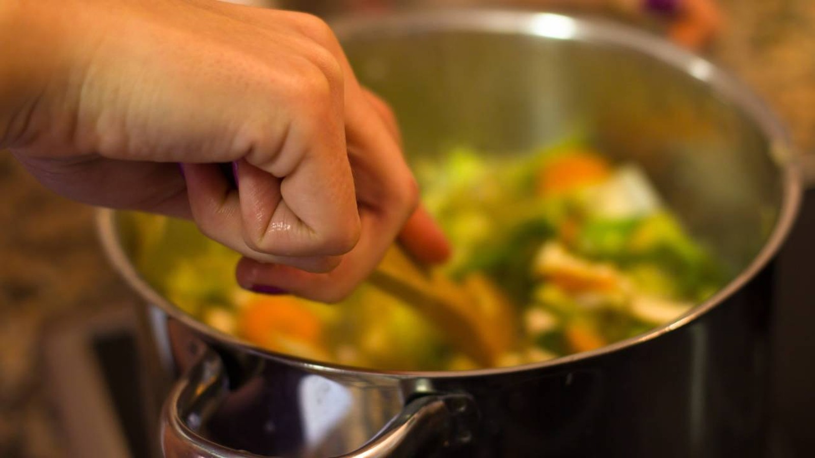 Mão mexendo legumes coloridos com colher de pau em panela no fogão