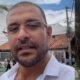 Homem com barba e óculos, camisa branca, fala à câmera em rua com prédios e árvore ao fundo