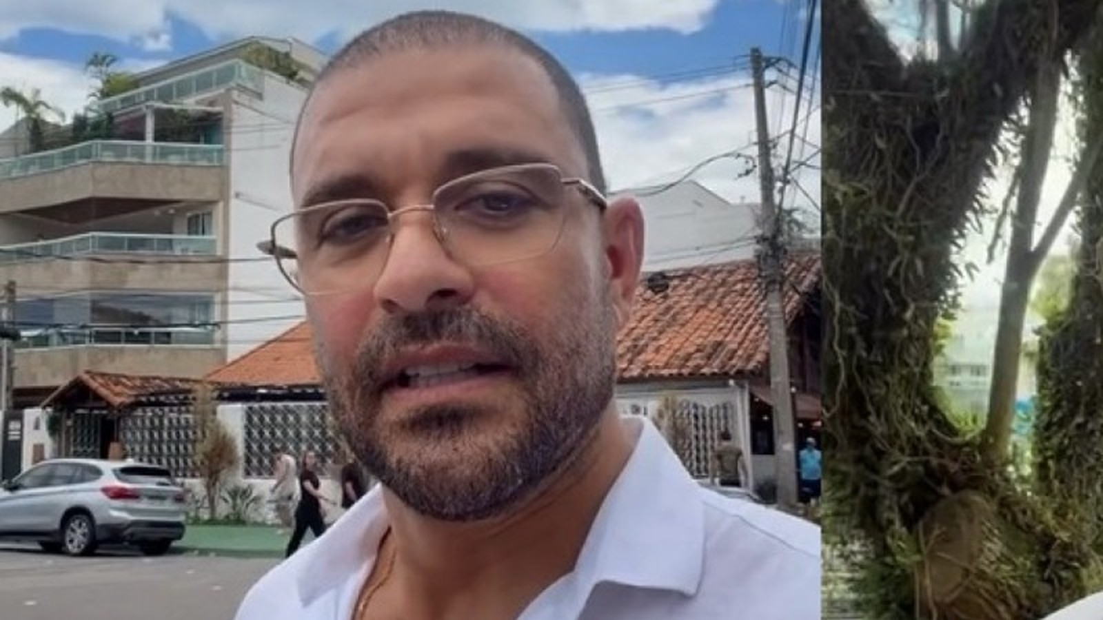 Homem com barba e óculos, camisa branca, fala à câmera em rua com prédios e árvore ao fundo