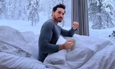 Homem barbudo em camisa de manga longa sentado na cama, segurando caneca, com paisagem nevada