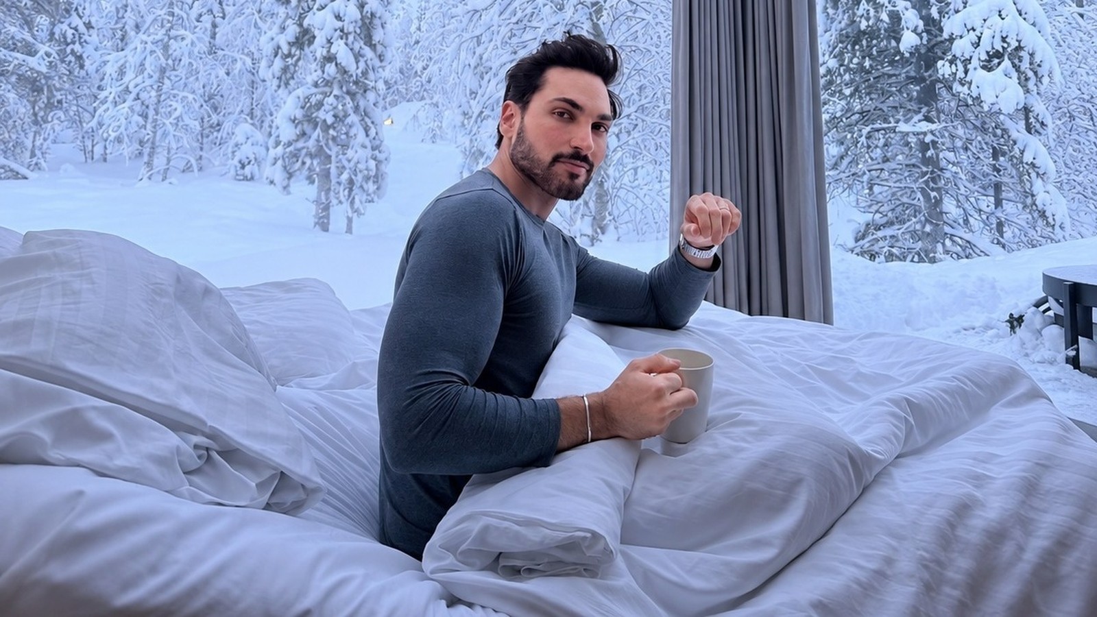 Homem barbudo em camisa de manga longa sentado na cama, segurando caneca, com paisagem nevada