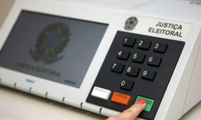 Mão pressiona botão "CONFIRMA" em urna eletrônica brasileira com teclado numérico visível