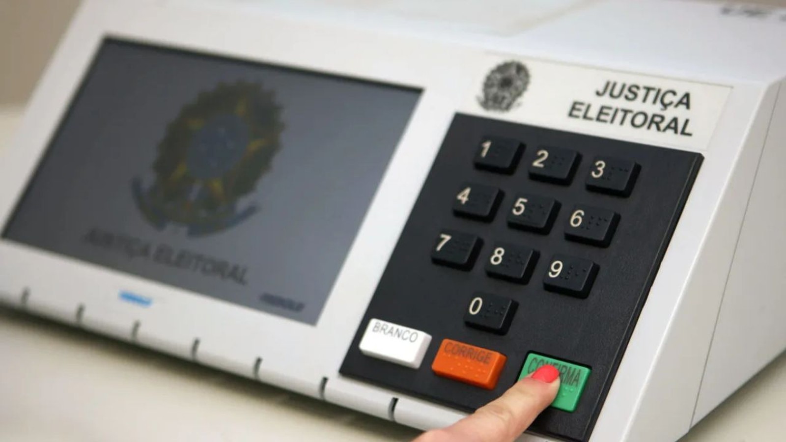 Mão pressiona botão "CONFIRMA" em urna eletrônica brasileira com teclado numérico visível
