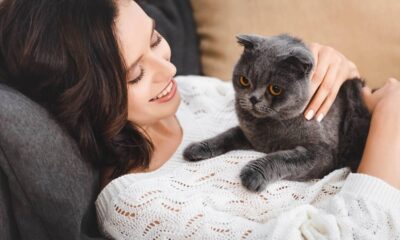 Mulher sorrindo, deitada com um gato cinza de olhos alaranjados descansando sobre ela