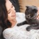 Mulher sorrindo, deitada com um gato cinza de olhos alaranjados descansando sobre ela