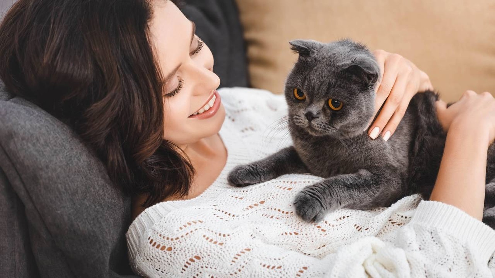 Mulher sorrindo, deitada com um gato cinza de olhos alaranjados descansando sobre ela