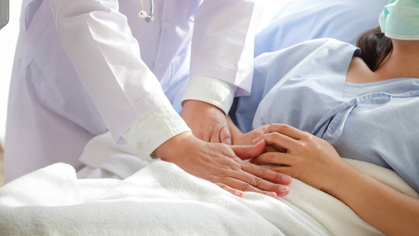 Mãos de médico examinando abdômen de paciente com máscara em leito hospitalar