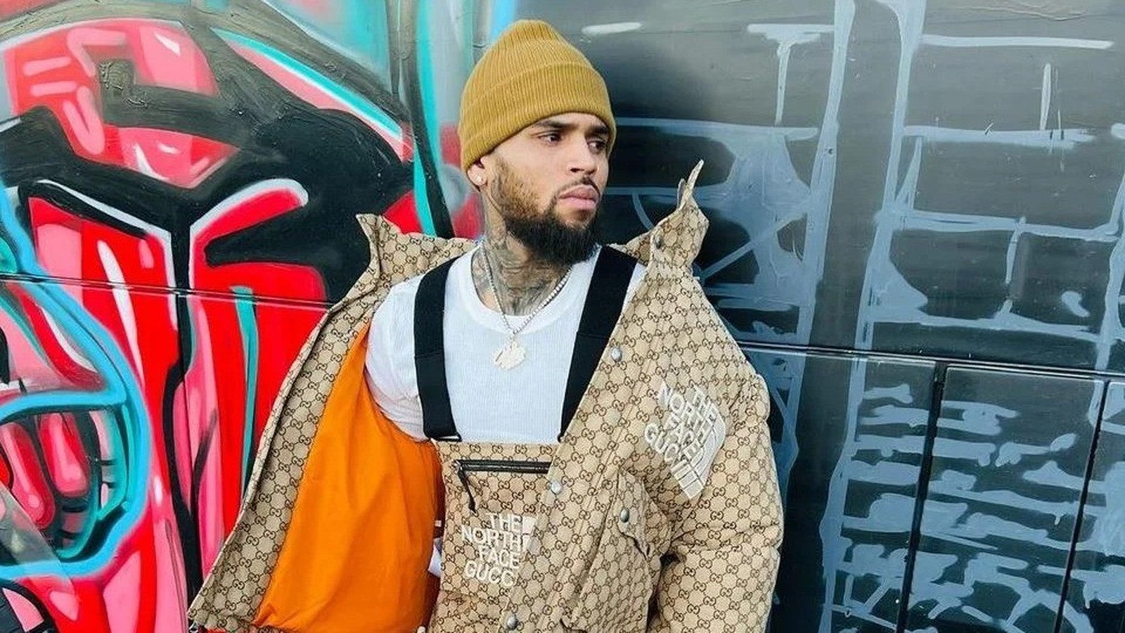 Chris Brown usa gorro amarelo, jaqueta The North Face Gucci, colar, contra parede grafite