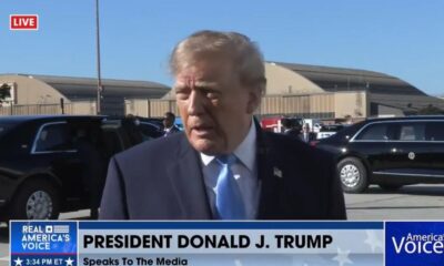 Donald Trump, terno azul, gravata azul, falando, com carros pretos, hangares e legendas de TV