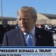 Donald Trump, terno azul, gravata azul, falando, com carros pretos, hangares e legendas de TV