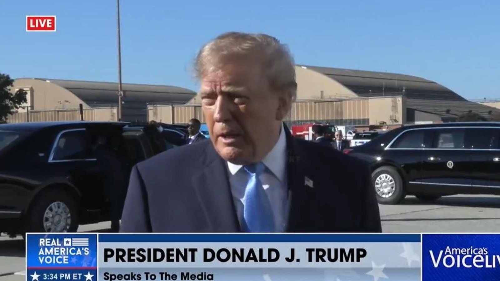 Donald Trump, terno azul, gravata azul, falando, com carros pretos, hangares e legendas de TV