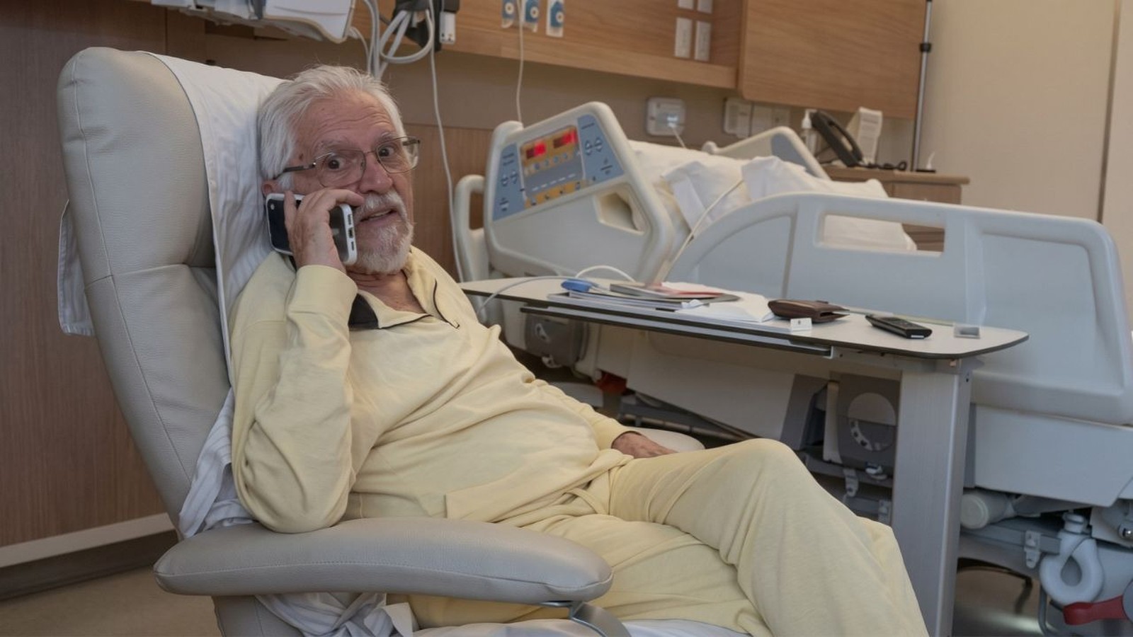Idoso de pijama amarelo, sentado em poltrona de hospital, falando ao celular