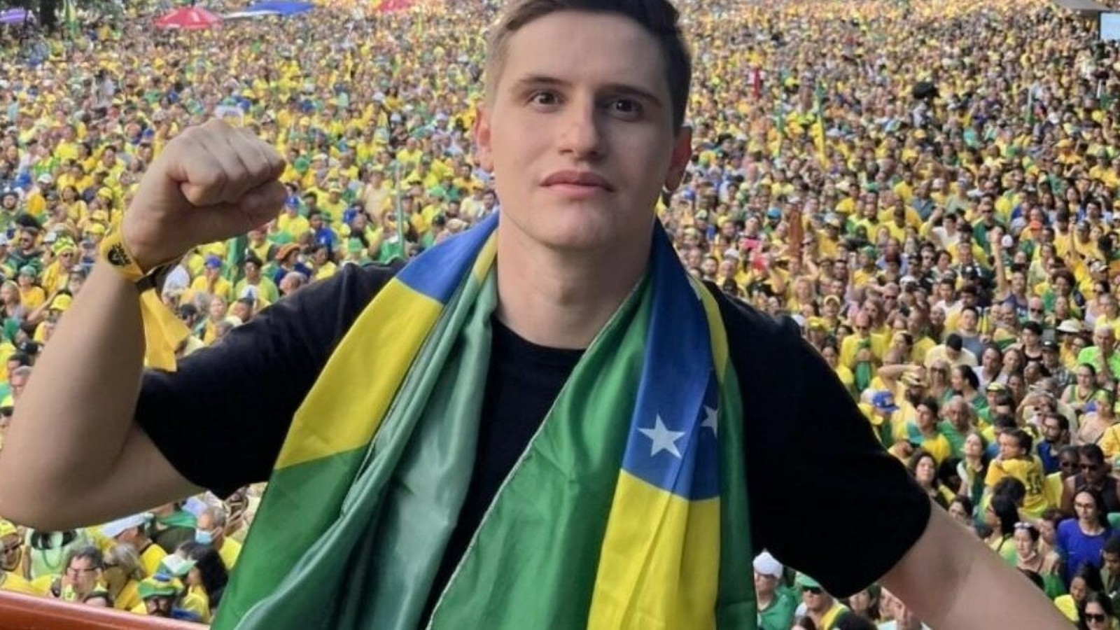 Homem com cachecol da bandeira do Brasil, punho cerrado, à frente de grande multidão