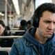 Homem com fones de ouvido, testa franzida, mão na têmpora, em um trem
