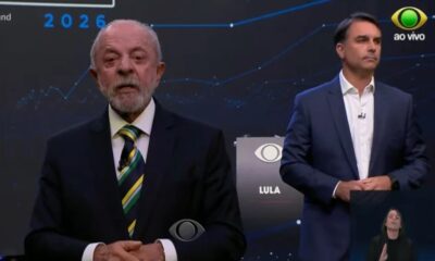 Lula fala em debate televisivo, Bolsonaro observa. Logos "Band", "ao vivo", "LULA", "2026" na tela