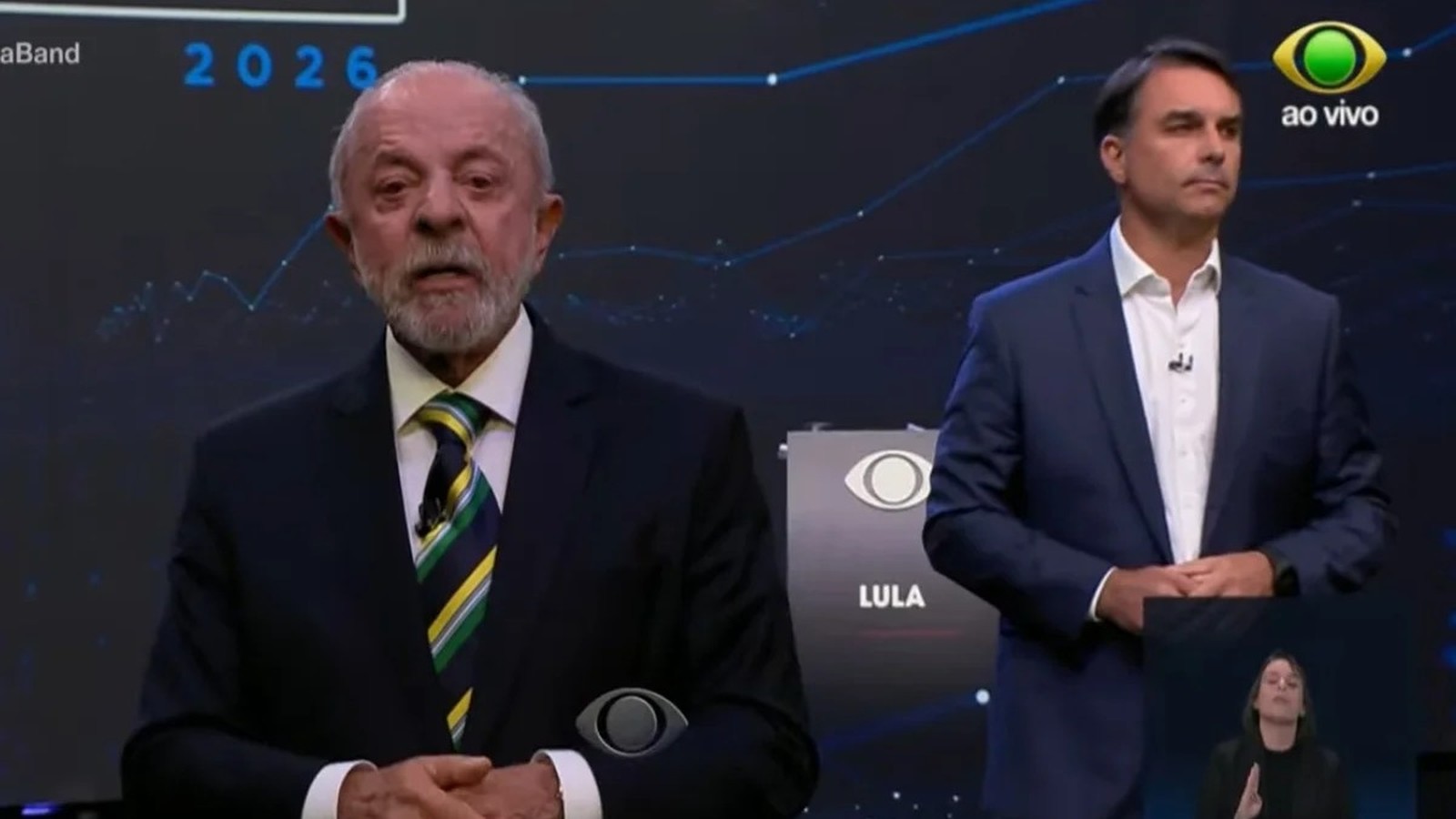 Lula fala em debate televisivo, Bolsonaro observa. Logos "Band", "ao vivo", "LULA", "2026" na tela