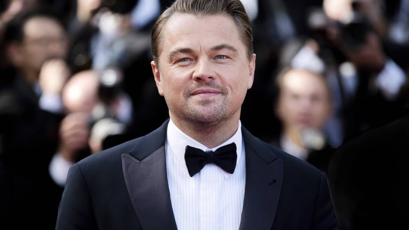 Leonardo DiCaprio em close-up, sorrindo levemente, vestindo terno preto e gravata borboleta