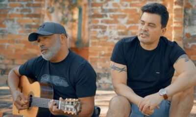 Homens cantam, um com violão e boné, outro com tatuagens, em ambiente externo