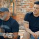 Homens cantam, um com violão e boné, outro com tatuagens, em ambiente externo