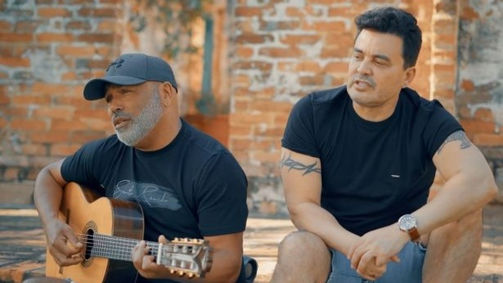 Homens cantam, um com violão e boné, outro com tatuagens, em ambiente externo