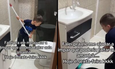 Menino pequeno de camiseta azul esfrega o chão do banheiro com um rodo próximo ao vaso e pia