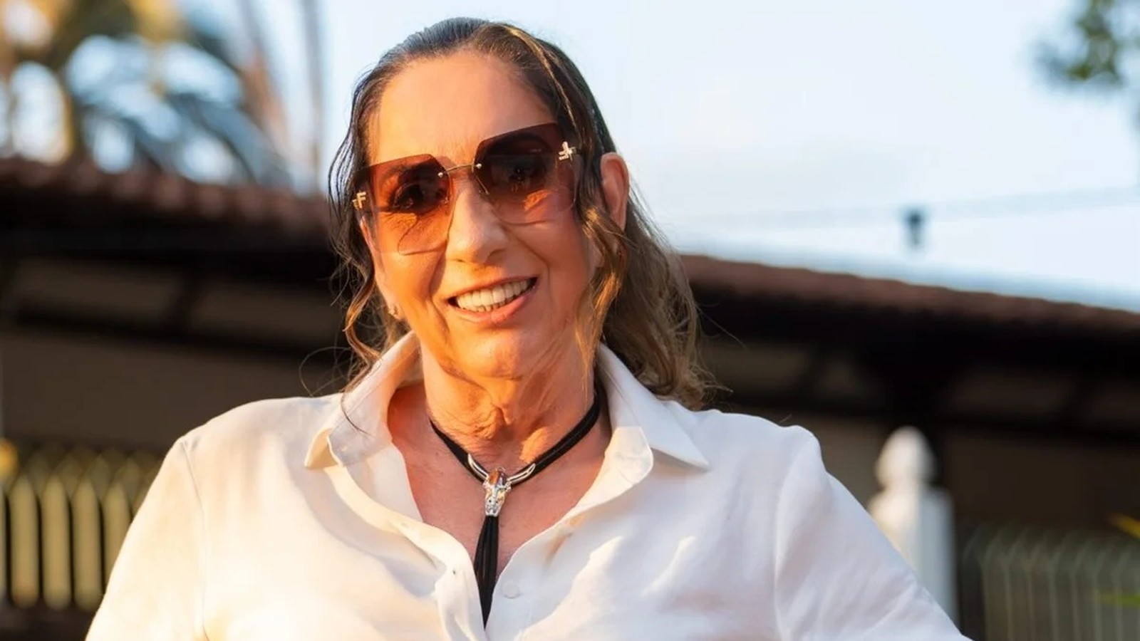 Margareth Serrão sorrindo, usando óculos escuros, camisa branca e colar com adorno prateado