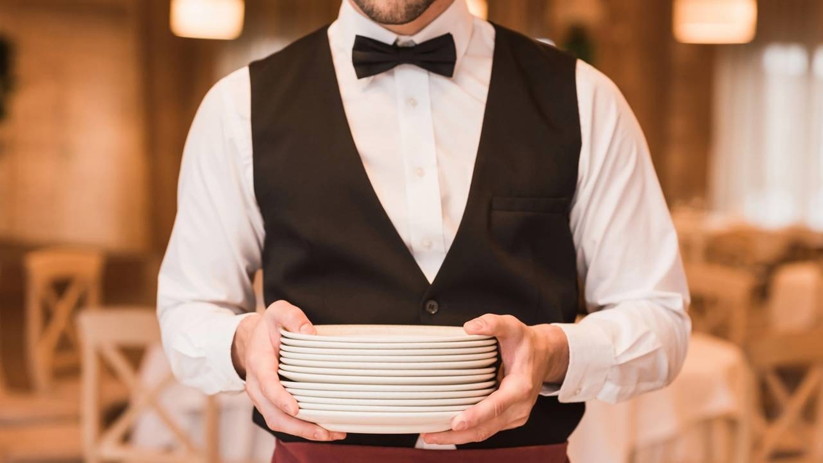 Homem de uniforme de garçom, colete preto, camisa branca, gravata borboleta, segurando pratos