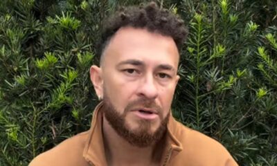 Homem com cabelo crespo, barba, jaqueta marrom e brinco, olhando para frente com fundo de arbustos