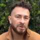 Homem com cabelo crespo, barba, jaqueta marrom e brinco, olhando para frente com fundo de arbustos