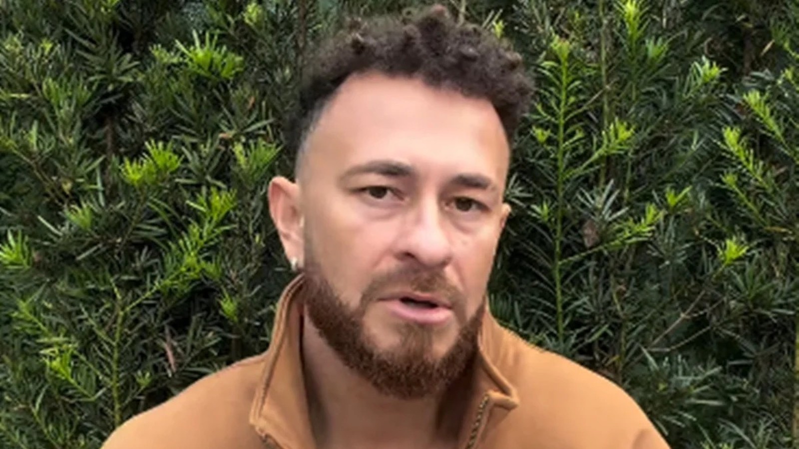 Homem com cabelo crespo, barba, jaqueta marrom e brinco, olhando para frente com fundo de arbustos
