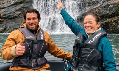 Duas pessoas sorrindo em um barco com coletes salva-vidas, cachoeira ao fundo