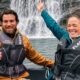Duas pessoas sorrindo em um barco com coletes salva-vidas, cachoeira ao fundo