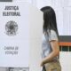 Mulher em cabine de votação com os dizeres "Justiça Eleitoral" e "Cabina de Votação"
