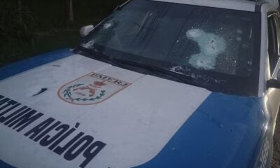 Viatura policial branca e azul com para-brisa e capô perfurados por tiros, logo PMERJ