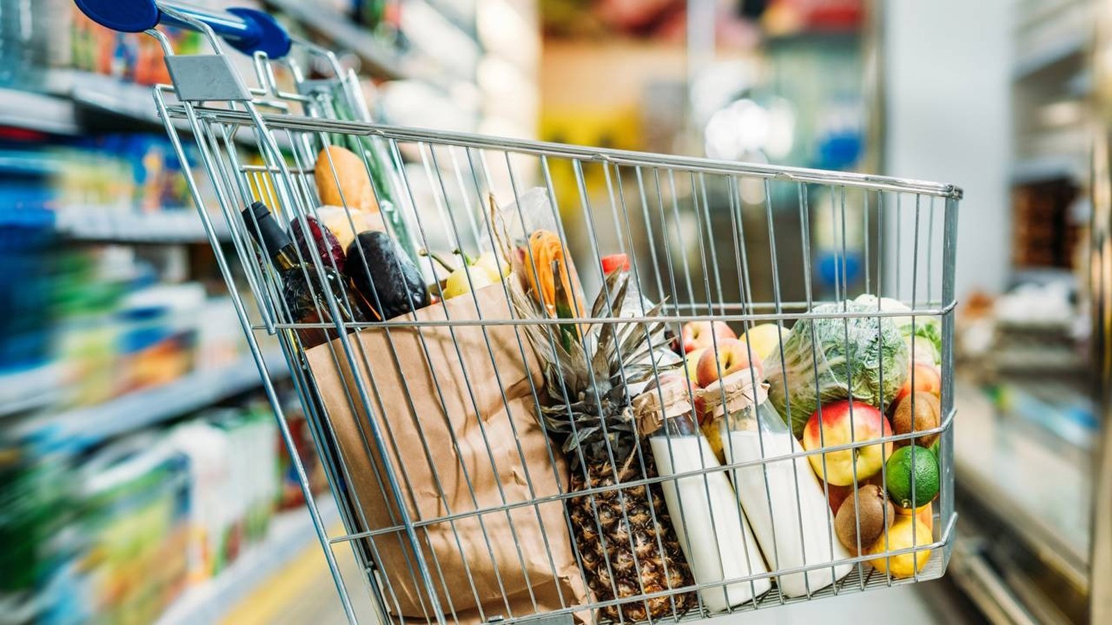 Carrinho de supermercado metálico cheio de frutas, vegetais, garrafas de leite e uma sacola de papel em corredor