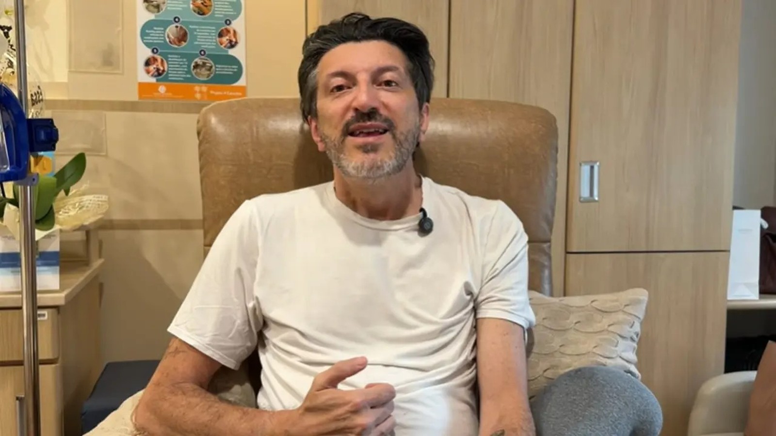 Lito Sousa, homem de barba e camiseta branca, sentado em poltrona, com microfone e IV.