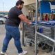 Homem forte devolve carrinho de compras; placa azul 'Devolva aqui seu carrinho' visível