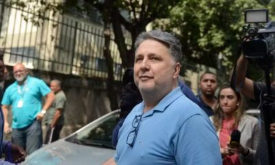Homem de polo azul com óculos, texto 'HI', rodeado por público e câmera de vídeo