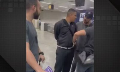Quatro homens em um aeroporto. Um homem de blusa preta é contido por outros dois