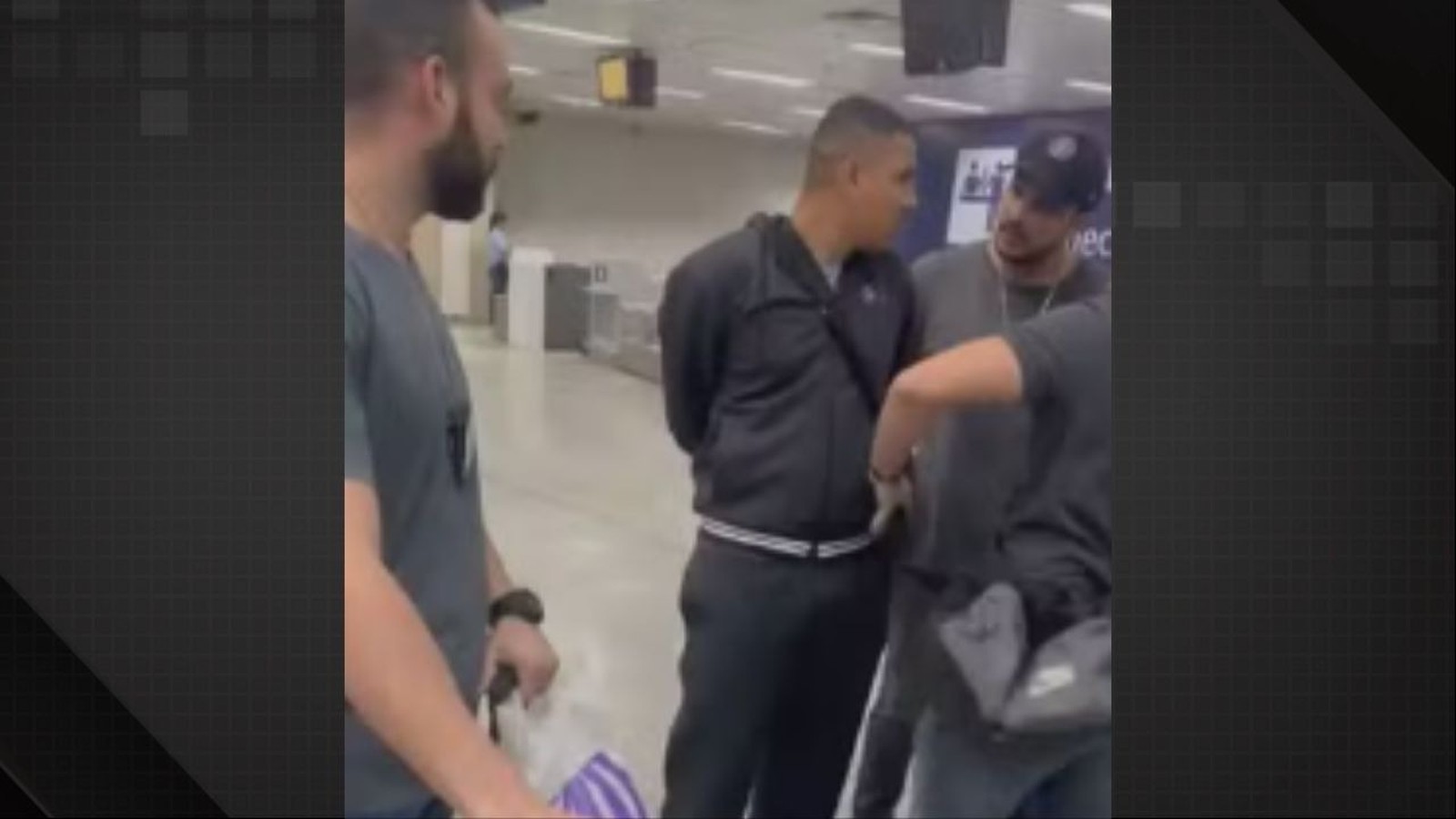 Quatro homens em um aeroporto. Um homem de blusa preta é contido por outros dois