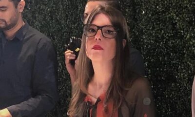 Mulher com óculos, cabelo castanho e batom vermelho olhando para a câmera, fundo verde