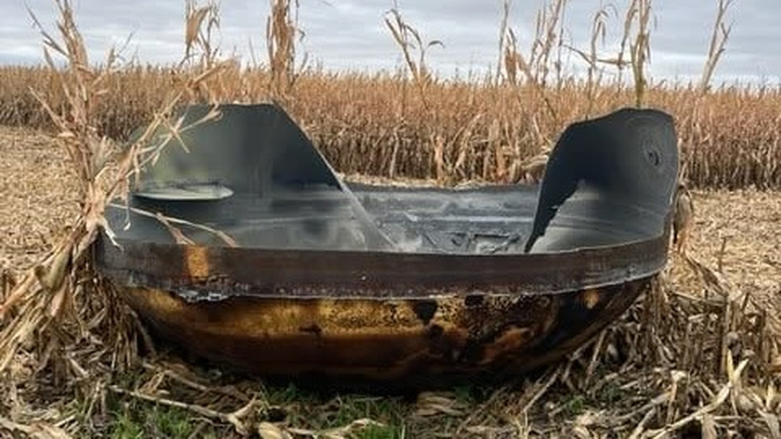 Grande destroço metálico escuro com marcas de queimadura em campo de milho seco