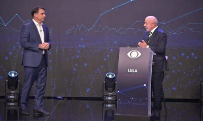 Dois homens em trajes formais em um palco de debate, com um pódio escrito "LULA"