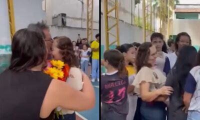 Duas mulheres se abraçam em quadra escolar. Alunos observam ao fundo. Camisa tem "ANA.LIGO 23" escrito